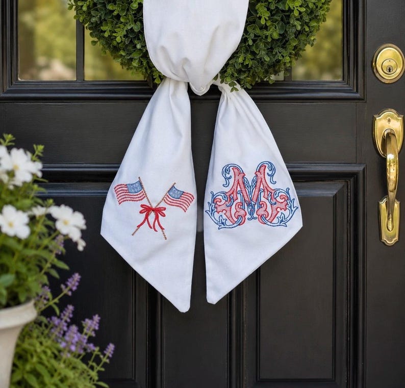 May include: A white fabric door hanger featuring two embroidered American flags crossed with a red bow on the left tail and a large, ornate, red and blue embroidered letter M on the right tail, draped over a green wreath on a black door.