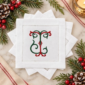May include: A stack of white square linen napkins featuring a hemstitched border and a central embroidered monogram in red and green thread. The design consists of a stylized letter T intertwined with a decorative scroll pattern in festive colors.