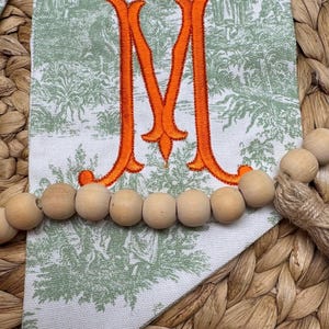 May include: A white fabric pennant featuring a green toile de jouy pattern and a large, embroidered orange letter M in the center. A strand of natural, light-colored wooden beads is draped horizontally across the front of the fabric pennant.