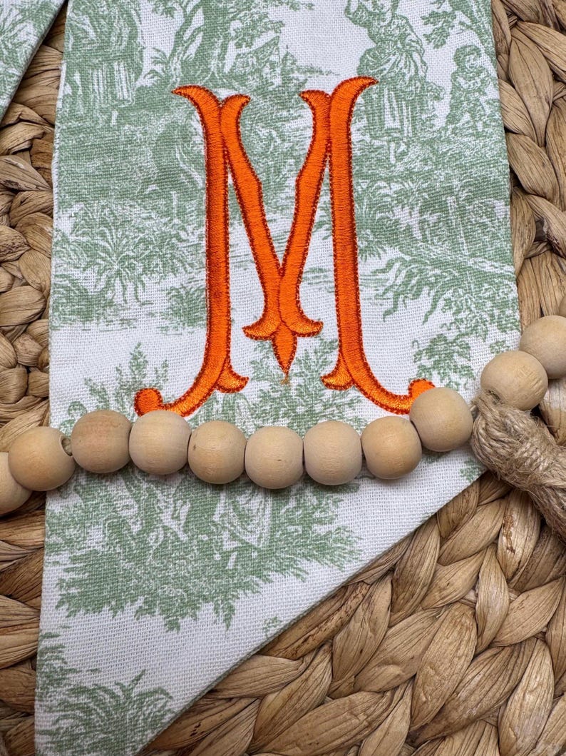 May include: A white fabric pennant featuring a green toile de jouy pattern and a large, embroidered orange letter M in the center. A strand of natural, light-colored wooden beads is draped horizontally across the front of the fabric pennant.