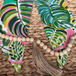 May include: A garland of wooden beads with tassels hangs on a woven wicker surface. The garland is decorated with pink and green fabric with a tropical leaf pattern.