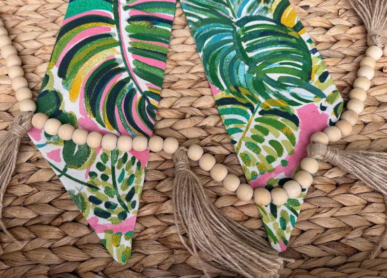 May include: A garland of wooden beads with tassels hangs on a woven wicker surface. The garland is decorated with pink and green fabric with a tropical leaf pattern.
