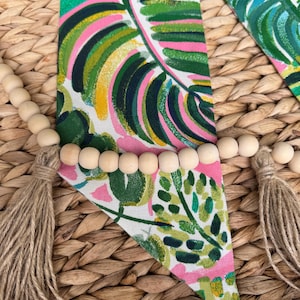 May include: A triangular fabric banner with a tropical leaf pattern in shades of green, pink, and yellow. The banner is decorated with a strand of wooden beads and two tassels.