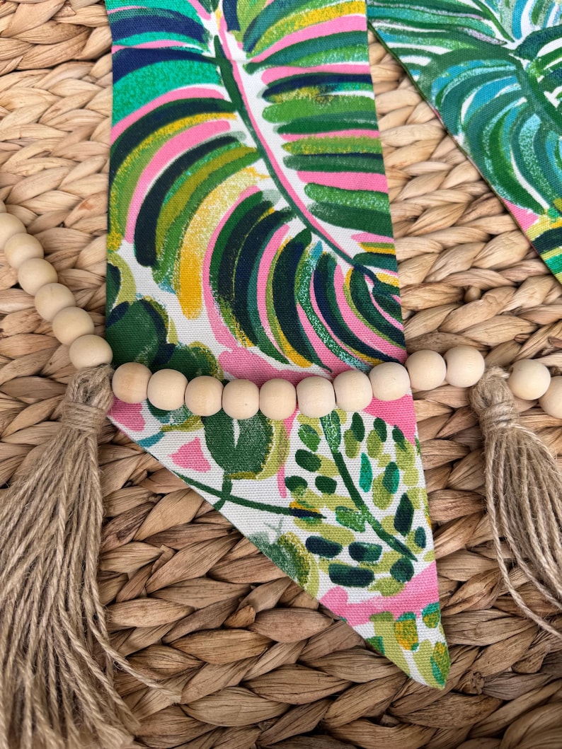 May include: A triangular fabric banner with a tropical leaf pattern in shades of green, pink, and yellow. The banner is decorated with a strand of wooden beads and two tassels.