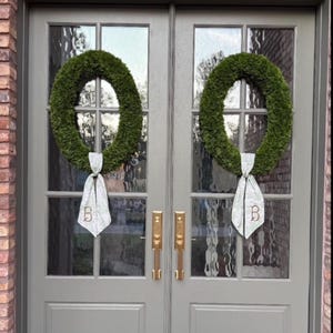 May include: Two oval-shaped green wreaths are hung on a set of double gray doors with glass panels. Each wreath features a light-colored fabric ribbon tied at the bottom, with the letter B embroidered in a dark thread on each ribbon tail.