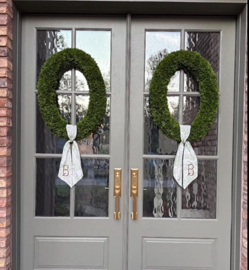 May include: Two oval-shaped green wreaths are hung on a set of double gray doors with glass panels. Each wreath features a light-colored fabric ribbon tied at the bottom, with the letter B embroidered in a dark thread on each ribbon tail.