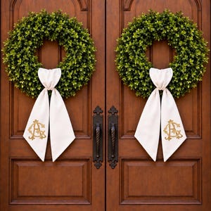May include: Two circular green boxwood wreaths are mounted on a pair of dark brown wooden doors. Each wreath features a white fabric ribbon tied at the bottom, with a gold embroidered monogram featuring the letters A and G on the fabric.