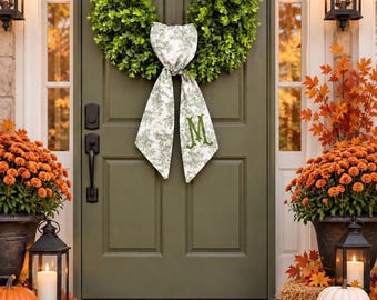 May include: A green boxwood wreath hangs on an olive green door, featuring a white and green toile fabric bow with a green embroidered letter M. The entryway includes orange chrysanthemums, pumpkins, lanterns, and autumn leaves on the porch.