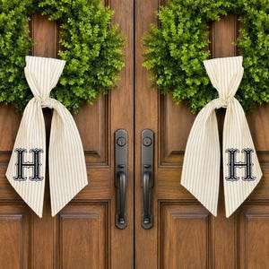 May include: Two circular green boxwood wreaths hanging on double wooden doors. Each wreath features a beige and white striped fabric ribbon tied in a bow, with the capital letter H embroidered in black on the lower portion of the ribbon tails.