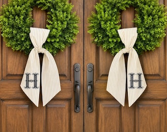 May include: Two circular green boxwood wreaths hanging on double wooden doors. Each wreath features a beige and white striped fabric ribbon tied in a bow, with the capital letter H embroidered in black on the lower portion of the ribbon tails.