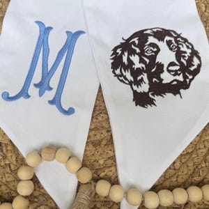 May include: Two white fabric pennants with blue embroidered letter "M" on one and a brown embroidered dog head on the other. The pennants are on a woven brown surface.