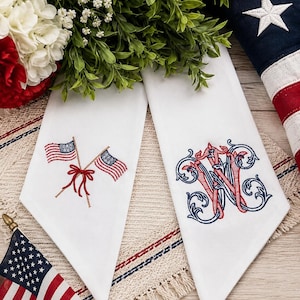 May include: Two white fabric napkins featuring detailed embroidery. One napkin displays two crossed American flags tied with a red bow. The other napkin features an ornate, intertwined monogram letter W in red and blue thread with scrollwork.