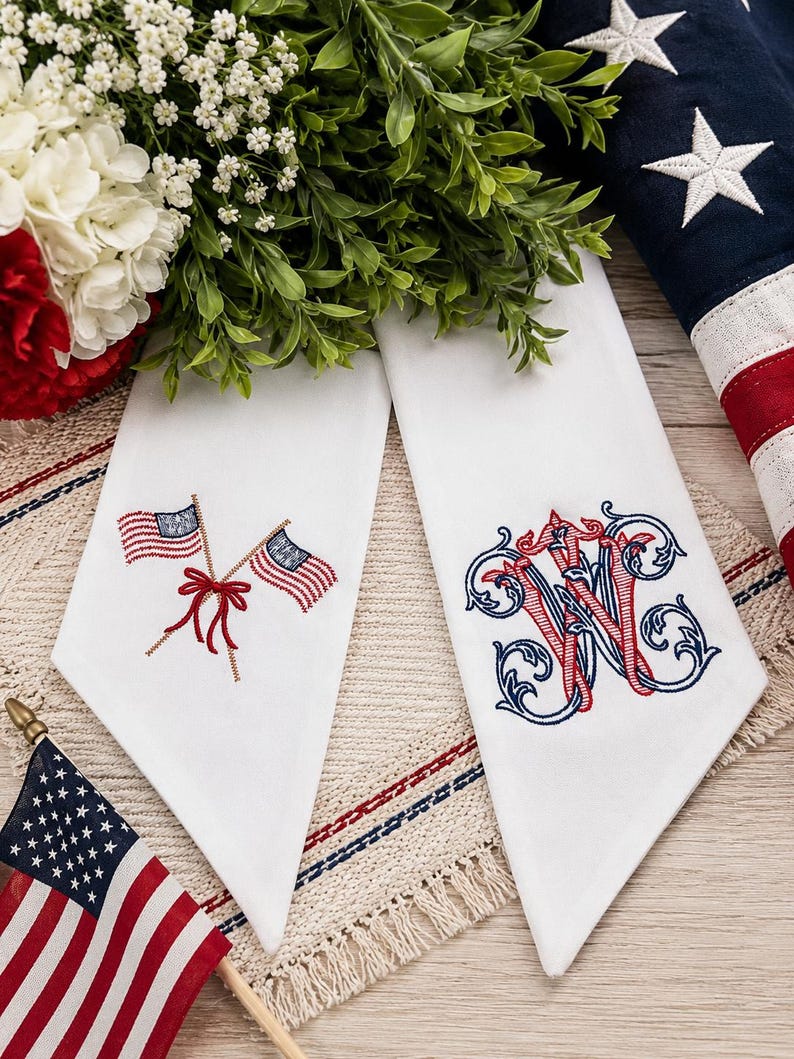 May include: Two white fabric napkins featuring detailed embroidery. One napkin displays two crossed American flags tied with a red bow. The other napkin features an ornate, intertwined monogram letter W in red and blue thread with scrollwork.