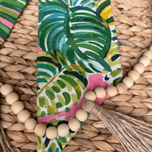 May include: A wooden bead garland with a natural jute tassel hangs on a woven wicker surface. The garland is draped over a pink and green fabric with a tropical leaf pattern.