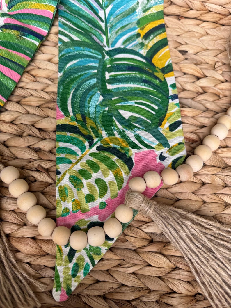 May include: A wooden bead garland with a natural jute tassel hangs on a woven wicker surface. The garland is draped over a pink and green fabric with a tropical leaf pattern.