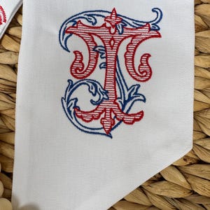 May include: A white fabric item featuring an embroidered monogram of the letter T in red thread with horizontal line details, surrounded by decorative blue scrollwork embroidery. The fabric has a visible woven texture and a pointed corner edge.