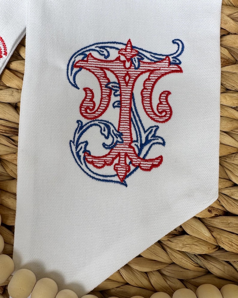 May include: A white fabric item featuring an embroidered monogram of the letter T in red thread with horizontal line details, surrounded by decorative blue scrollwork embroidery. The fabric has a visible woven texture and a pointed corner edge.
