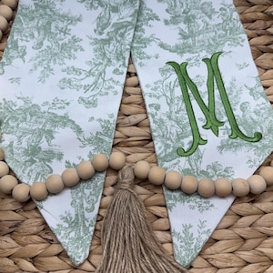 May include: Two fabric pennants featuring a sage green toile de jouy pattern on a white background. One pennant displays a large, embroidered sage green letter M. A string of natural wooden beads with a central jute tassel connects the two pieces.
