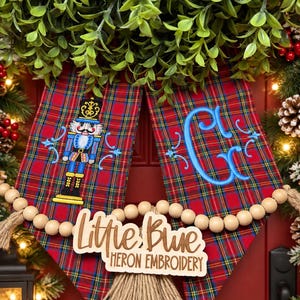 May include: A red tartan fabric banner featuring an embroidered blue nutcracker on the left and a blue monogram letter C on the right. A wooden bead garland hangs across the front with a wooden sign reading Little Blue Heron Embroidery.