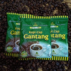 Könnte beinhalten: Drei grüne Kaffeepackungen mit gelbem Text und einer Kaffeetassen-Illustration sind von Kaffeebohnen umgeben. Auf den Packungen steht "Kopi Cap Gantang" und "SEJAK 1990".