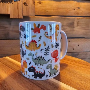 Può includere: Tazza in ceramica bianca con un motivo di dinosauri colorati. Il design presenta vari dinosauri dei cartoni animati in tonalità arancioni, gialle, verdi e nere, insieme ad accenti di foglie verdi. La tazza ha un manico bianco ed è appoggiata su una superficie di legno.