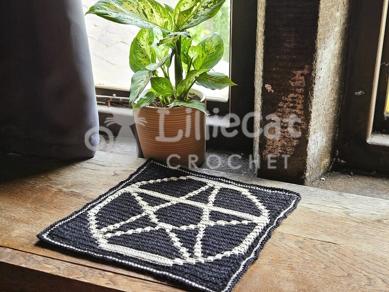 A crochet pattern using overlay mosaic crochet technique. The pattern has a dark background with a white pentagram motif in the middle. The photo is taken in a window with a pot plant behind. The photo has an alternative and witchy feel.