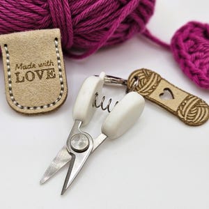 May include: A set of knitting tools including a white spring-loaded scissor, a brown leather tag with the text "Made with LOVE", and a brown wooden charm with a heart cutout and a yarn ball design.
