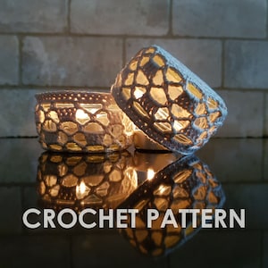May include: Two glass candle holders with a crocheted white overlay, illuminated by candlelight. The crochet pattern creates a lattice effect, and the text "CROCHET PATTERN" is displayed at the bottom. The warm glow of the candles is reflected on the surface.