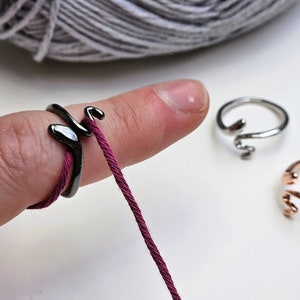 May include: A black metal ring with a hook for holding yarn. The ring is worn on a finger and has a burgundy yarn threaded through the hook.