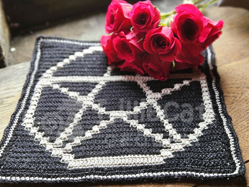 A crochet pattern using overlay mosaic crochet technique. The pattern has a dark background with a white pentagram motif in the middle. The photo is taken in a window and there are red roses. The photo has an alternative and witchy feel.