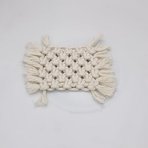 May include: A square, off-white macrame coaster with a woven pattern and fringe detailing. The coaster is made of thick, natural-colored cotton cord, and is designed to protect surfaces from heat and moisture.