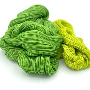 May include: Two skeins of yarn, one green and one yellow, are piled together. The yarn is soft and appears to be made of cotton.