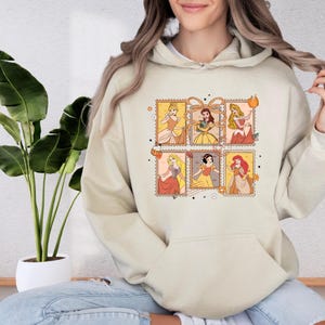A woman wearing a beige hoodie with a graphic of Disney princesses on it, sitting in front of a potted plant.