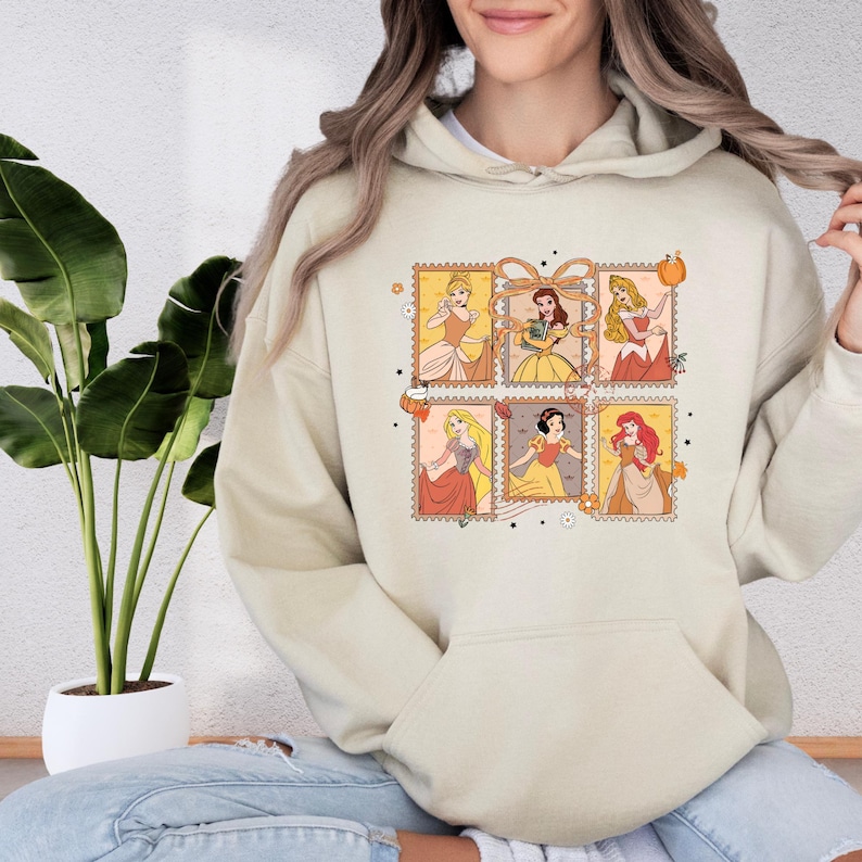 A woman wearing a beige hoodie with a graphic of Disney princesses on it, sitting in front of a potted plant.
