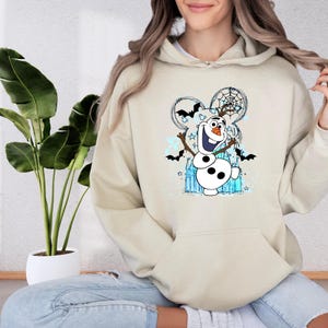 A woman wearing a beige hoodie with a graphic of a snowman and bats on it, sitting in front of a potted plant.