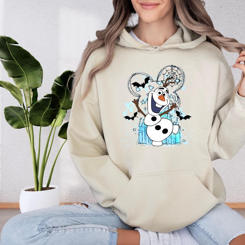 A woman wearing a beige hoodie with a graphic of a snowman and bats on it, sitting in front of a potted plant.