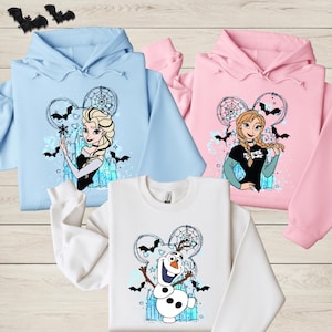 three hoodies with cartoon characters from the Disney movie Frozen. The hoodies are arranged on a wooden surface, with bats and a dreamcatcher design visible.