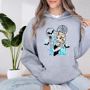 A woman wearing a grey hoodie with a graphic of a cartoon character, specifically Elsa from Frozen, is sitting on the floor next to a potted plant.