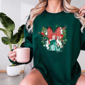 A woman wearing a green sweater with a graphic of a mermaid holding a mug, with a potted plant in the background.