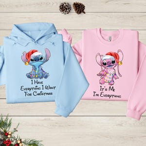 two sweatshirts with a cartoon character from the Disney movie Lilo and Stitch. The sweatshirt on the left is blue and the one on the right is pink. Both sweatshirts have a message printed on them, expressing the sentiment that the viewer has everything they want for Christmas.