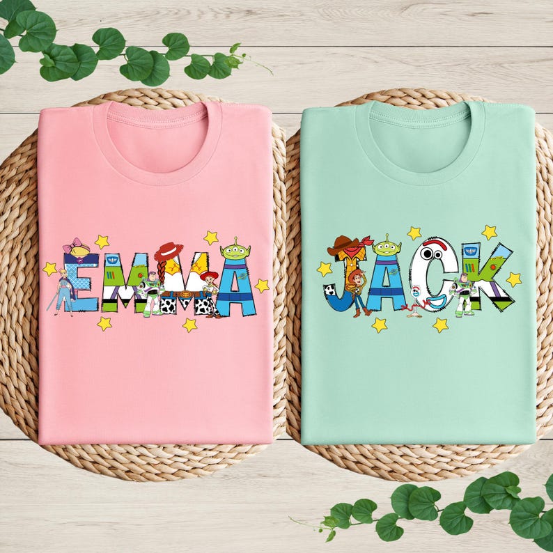 May include: Two t-shirts, one pink and one mint green, each with a name design. The pink shirt says "EMMA" in colourful letters with cartoon character accents. The mint green shirt says "JACK" with similar cartoon accents and star details.