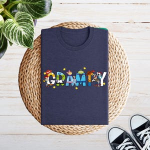 May include: A navy blue t-shirt featuring the word "Grammy" in colourful letters, each filled with cartoon characters and yellow stars. The shirt is folded on a woven mat, with black and white trainers in the corner.