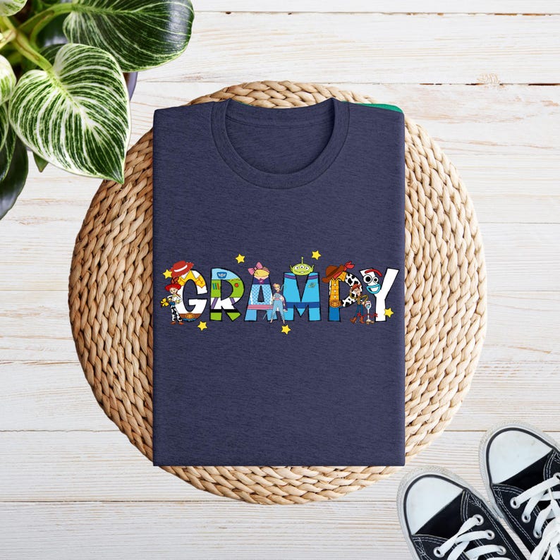 May include: A navy blue t-shirt featuring the word "Grammy" in colourful letters, each filled with cartoon characters and yellow stars. The shirt is folded on a woven mat, with black and white trainers in the corner.