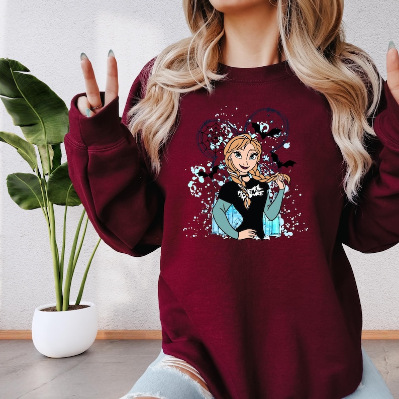 A woman wearing a burgundy sweatshirt with a graphic of a cartoon character on it, sitting in front of a potted plant.