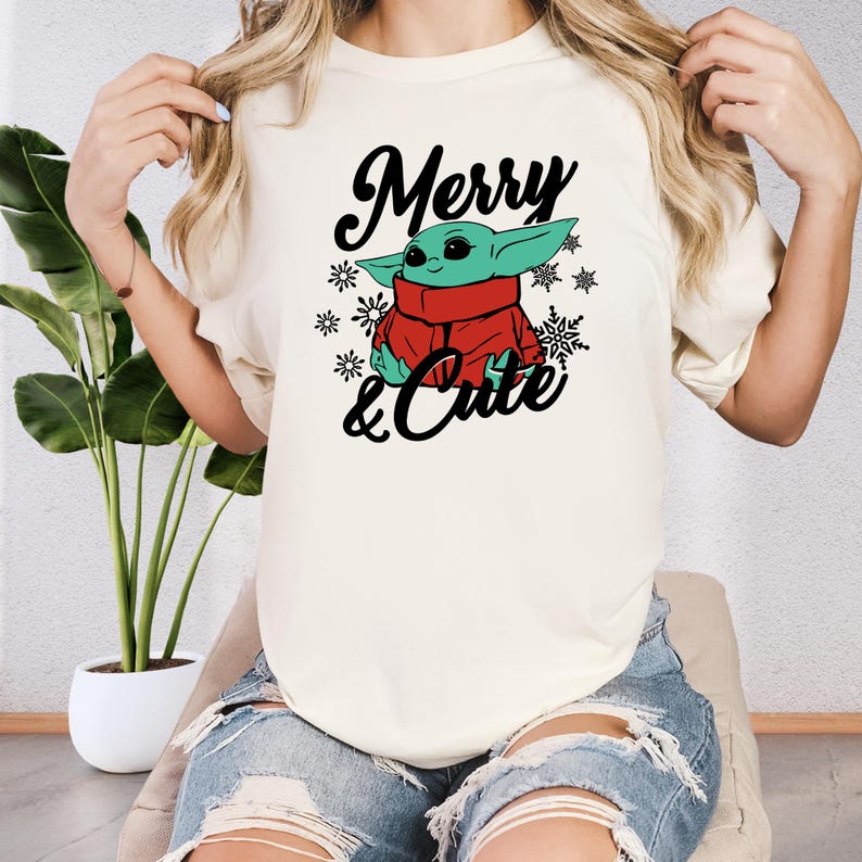 A woman wearing a white t-shirt with a graphic design of a young child wearing a red scarf and the text "Merry & Cute" printed on it.