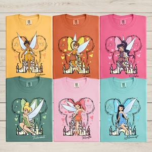 May include: Six colorful t-shirts featuring fairy characters. Each shirt displays a different fairy design with a castle and a Mickey Mouse-shaped outline. The shirts are in shades of yellow, orange, pink, green, and blue.