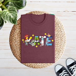 May include: A maroon t-shirt featuring the name "KATE" in a colourful, cartoon-themed design. The letters are adorned with characters and yellow stars. The shirt is folded on a woven mat, with black trainers visible.