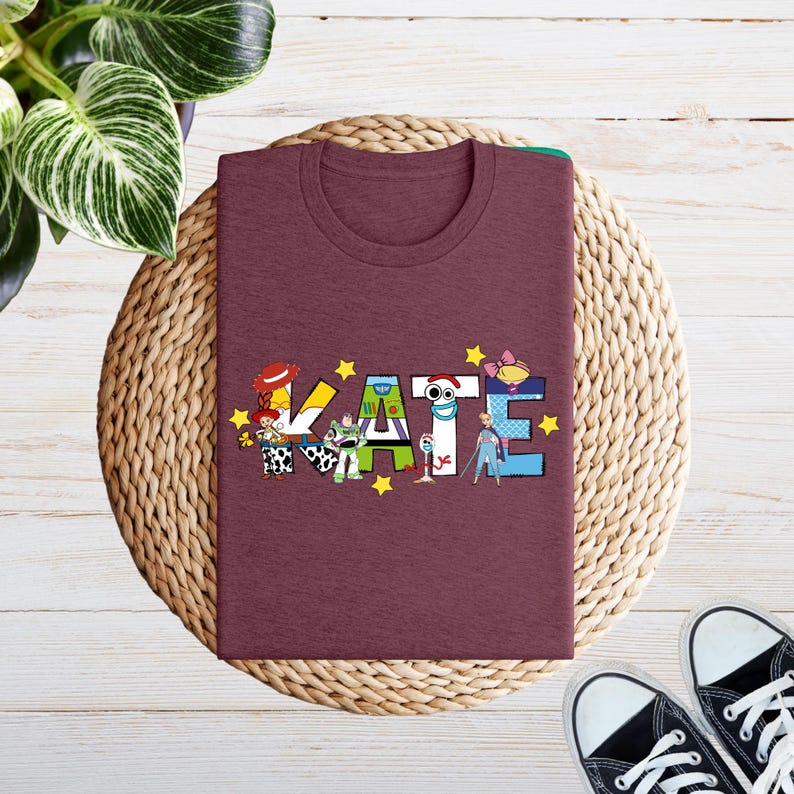 May include: A maroon t-shirt featuring the name "KATE" in a colourful, cartoon-themed design. The letters are adorned with characters and yellow stars. The shirt is folded on a woven mat, with black trainers visible.