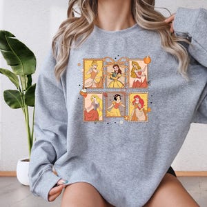 A woman wearing a gray sweatshirt with a graphic design featuring illustrations of Disney princesses.