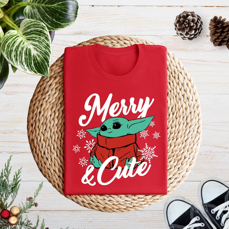 A red t-shirt with a graphic of a young alien character, likely representing the character Baby Yoda from the Star Wars franchise, is displayed on a woven placemat. The t-shirt has the text "Merry & Cute" printed on it. The image also includes pine cones and a pair of sneakers, suggesting a holiday or seasonal setting.